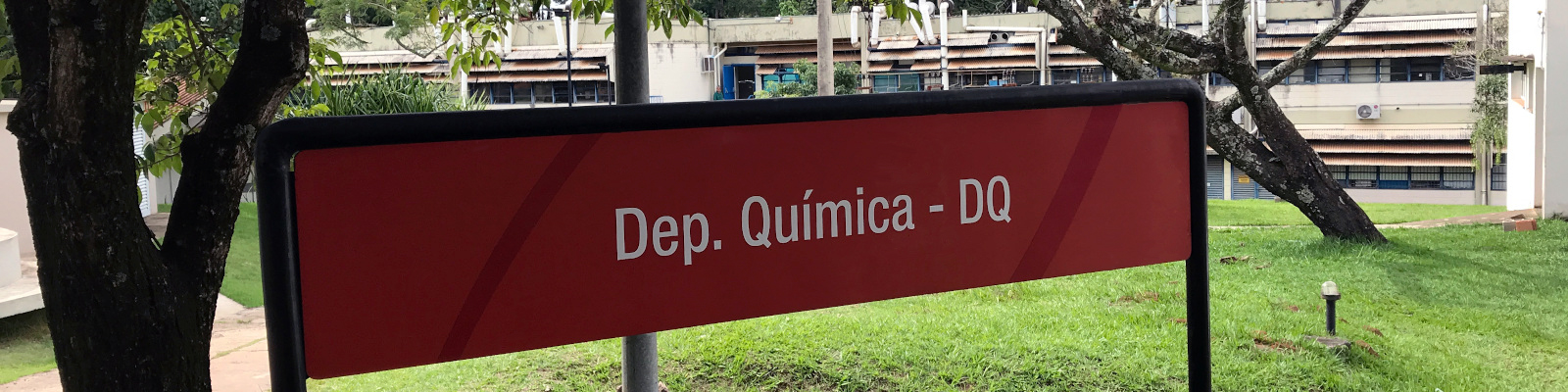 The image shows, in the foreground, a red sign, taking up most of the image, with white writing “Dep. Química – DQ” (Chemistry Dept.), serving to identify where the department is located. In the background, it is possible to notice the northern façade of the department, which is white, with several windows and apparent installations, in addition to some of the local vegetation.