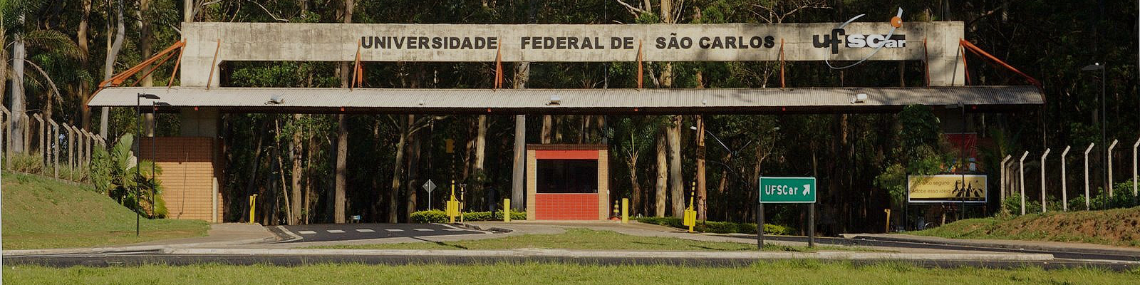 The image represents the entrance to the southern area of the UFSCar campus in São Carlos. The entrance has a guard post in the center and vehicle circulation to the left and right, in addition to the black sign written “Universidade Federal de São Carlos”(Federal University of São Carlos). The vegetation appears in the background, and it is possible to notice several trees, which comprise a good part of the campus landscape.